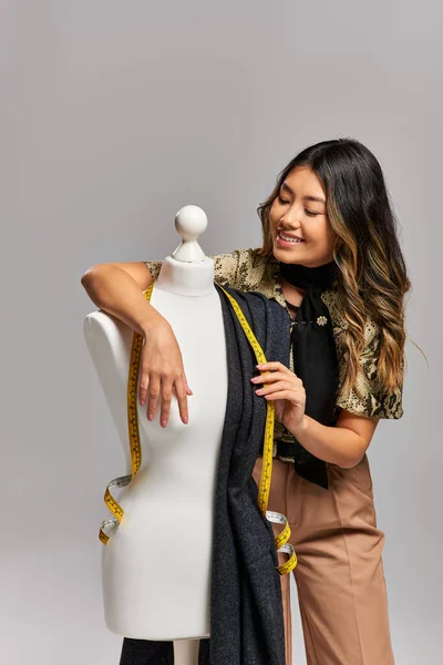 Pleased asian designer posing near mannequin with fabric and measuring tape on grey backdrop — Stock Photo
