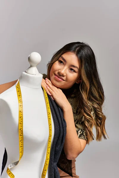 Dreamy asian clothes designer looking away near mannequin with measuring tape on grey backdrop — Stock Photo