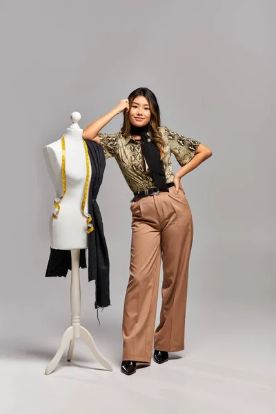 Модный азиатский модельер позирует рядом с манекеном с измерительной лентой и тканью на сером фоне — стоковое фото