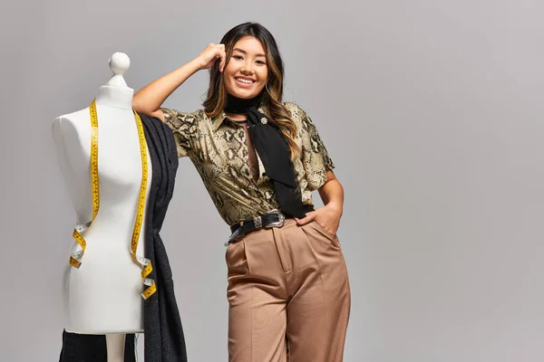 Fröhliche asiatische Schneiderin blickt in die Kamera neben Schaufensterpuppe mit Maßband und Stoff auf grau — Stockfoto
