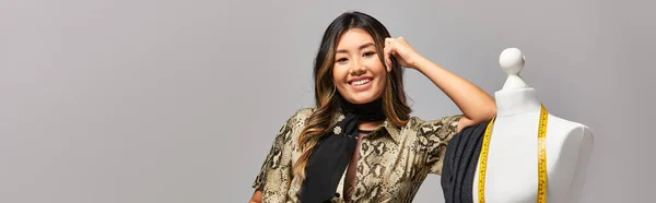 Joyful asian dressmaker smiling at camera near mannequin with measuring tape on grey, banner — Stock Photo