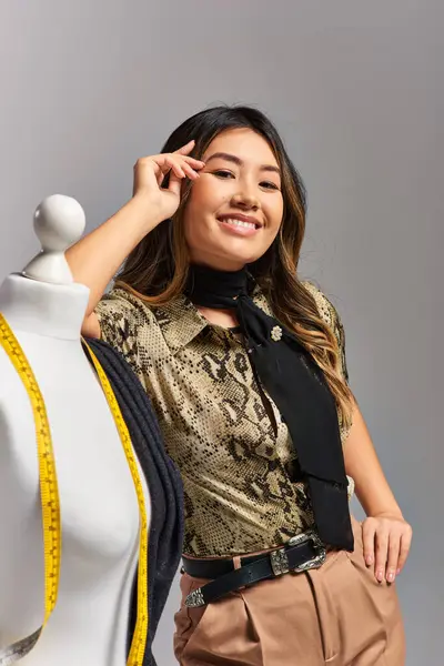 Smiling asian fashion designer with measuring tape near mannequin with fabric on grey backdrop — Stock Photo
