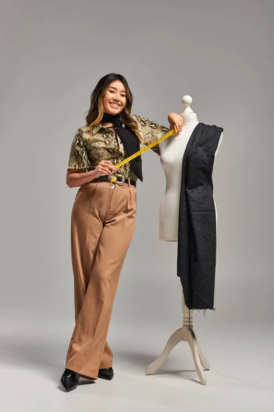 Young asian clothes designer holding measuring tape near mannequin with fabric on grey backdrop — Stock Photo