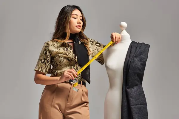 Весёлый азиатский стилист держит измерительную ленту рядом с манекеном с тканью на сером фоне — стоковое фото