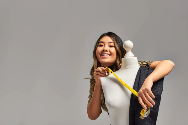 Positive asian designer holding measuring tape near mannequin with fabric on grey backdrop — Stock Photo