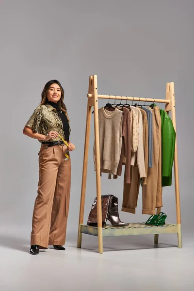 Creativo asiatico designer con metro nastro vicino rack con abiti su misura su sfondo grigio — Foto stock