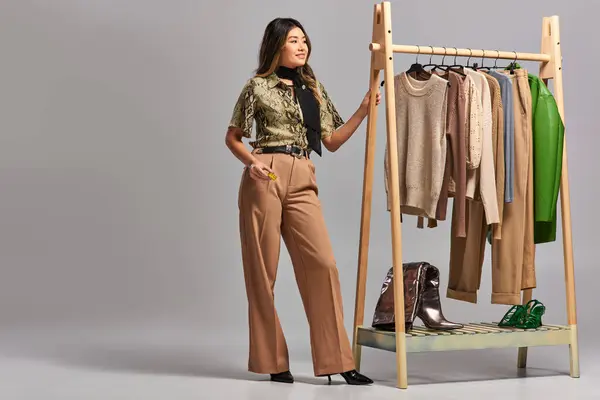 Happy asian designer with measuring tape looking away near rack with stylish clothes on grey — Stock Photo