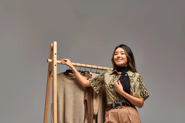 Young happy asian clothes stylist posing near rack with trendy bespoke clothes on grey backdrop — Stock Photo