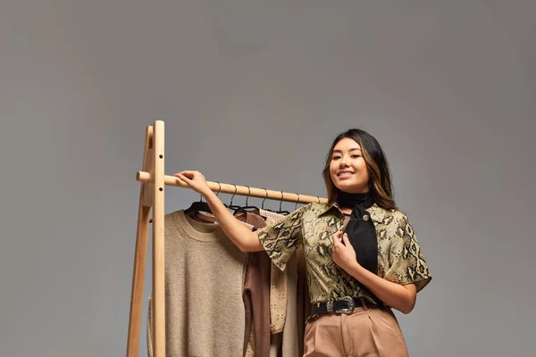 Jeune heureux asiatique vêtements styliste posant près de rack avec tendances vêtements sur mesure sur fond gris — Photo de stock