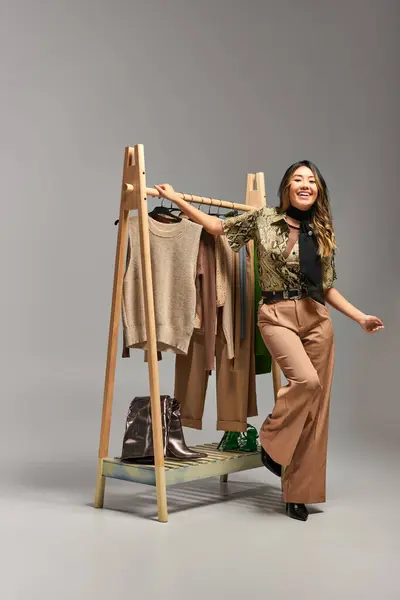 Overjoyed asian stylist smiling near rack with bespoke clothes on grey backdrop, fashion business — Stock Photo