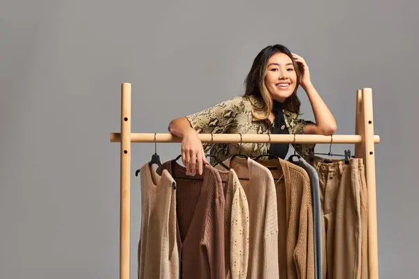 Cheerful asian stylist smiling at camera near rack with bespoke clothes on grey, fashion business — Stock Photo