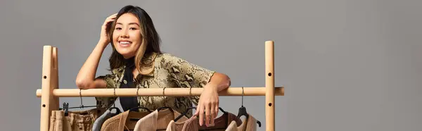 Довольный азиатский дизайнер позирует возле стойки с модной на заказ одеждой на сером, горизонтальном баннере — стоковое фото