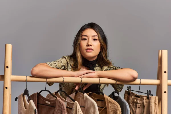 Selbstbewusste erfolgreiche asiatische Modedesignerin blickt mit Kleidungsstücken auf grau in die Kamera — Stockfoto