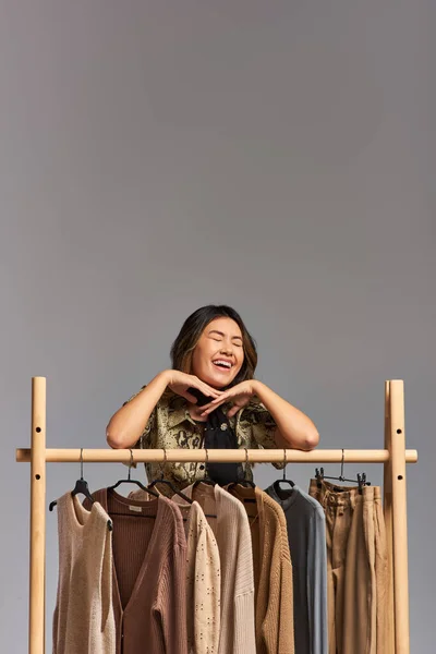 Взволнованный азиатский дизайнер одежды смеется с закрытыми глазами возле стойки с модной одеждой на сером — стоковое фото