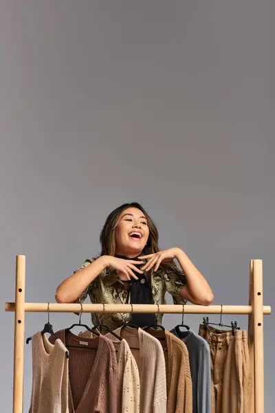Задоволений азіатський стиліст одягу сміється біля стійки з модним одягом на сірому тлі — стокове фото