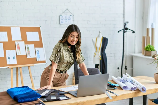 Young smiling asian clothes designer looking at laptop on work desk in personal fashion atelier — Stock Photo