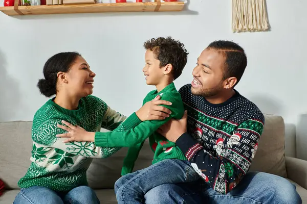 Familia afroamericana feliz sentado en el sofá y sonriendo alegremente el uno al otro, Navidad - foto de stock