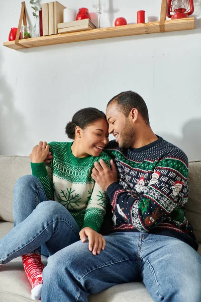 Couple amoureux afro-américain en chandails d'hiver étreignant chaudement avec les yeux fermés sur le canapé, Noël — Photo de stock