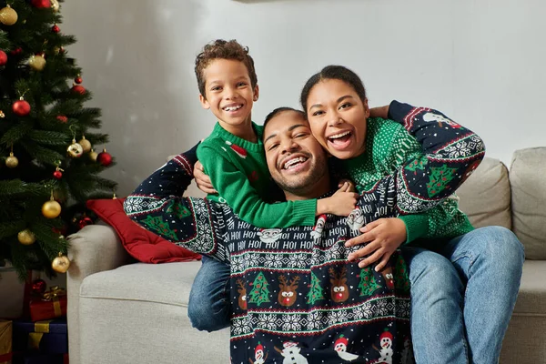 Familia afroamericana feliz en suéteres acogedores abrazándose calurosamente y sonriendo a la cámara, Navidad - foto de stock