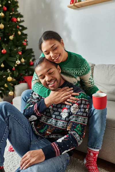 Tiro vertical de la feliz pareja afroamericana en suéteres cálidos abrazando con los ojos cerrados, Navidad - foto de stock
