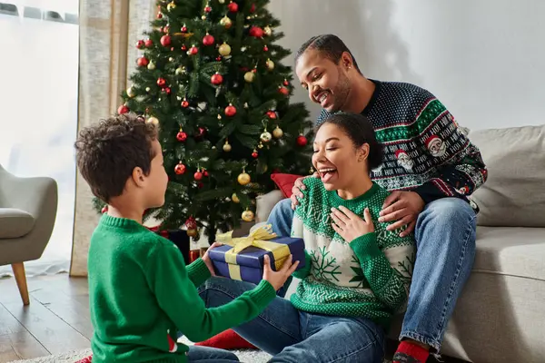 Adorabile ragazzo afroamericano che fa un regalo ai suoi genitori accanto all'albero di Natale con le bagattelle — Foto stock