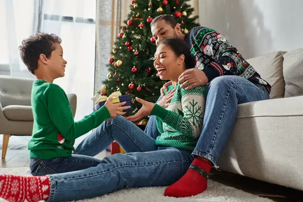 Mignon garçon afro-américain joyeux en pull décontracté chaud donnant cadeau à ses parents, Noël — Photo de stock