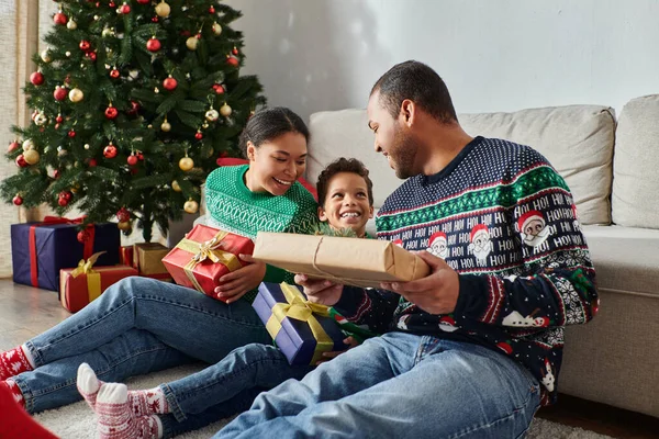 Feliz família afro-americana sentada no chão segurando presentes e sorrindo alegremente, Natal — Fotografia de Stock