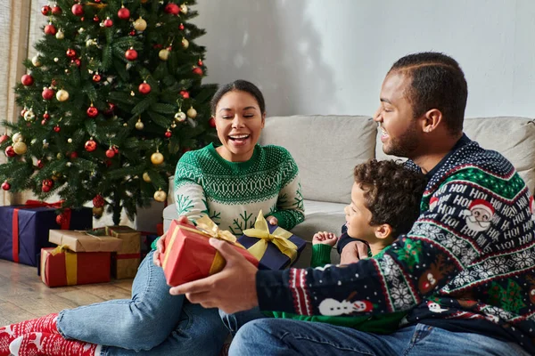 Heureuse belle famille en pull chaud échangeant des cadeaux et souriant joyeusement, Noël — Photo de stock