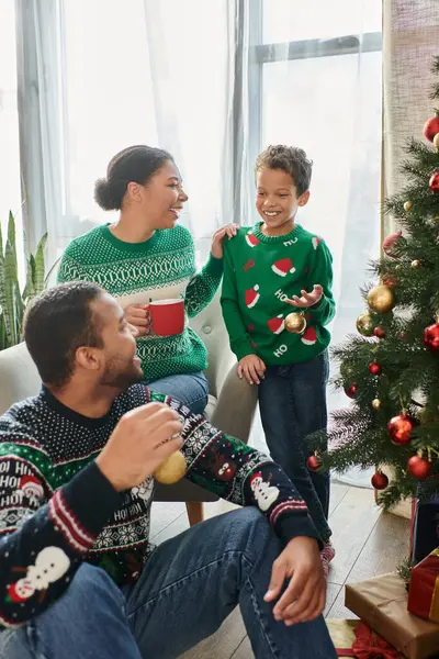 Mujer afroamericana con taza de café mirando a su marido y su hijo decorando el árbol de Navidad - foto de stock
