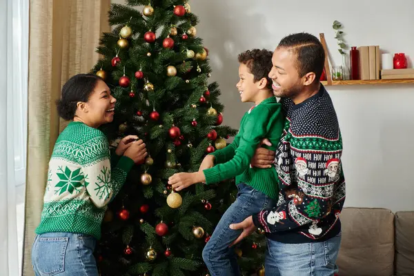Alegre afro-americana mãe olhando para o marido ajudando seu filho a decorar a árvore de Natal — Fotografia de Stock