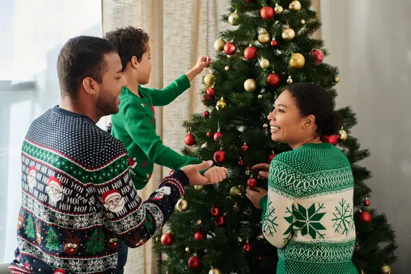 Веселые любящие африканские американские родители в теплых свитерах помогают своему сыну украсить елку — стоковое фото