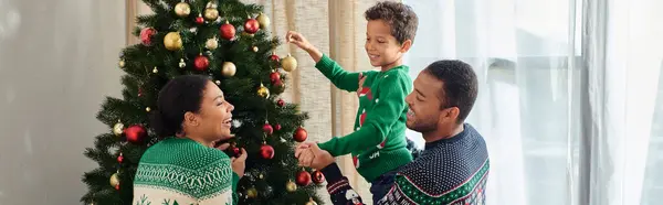 Cheerful african american family in warm sweaters putting baubles on Christmas tree together, banner — Stock Photo