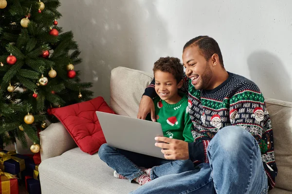 Feliz lindo afroamericano chico viendo la película de comedia de Navidad en el ordenador portátil con su padre alegre - foto de stock