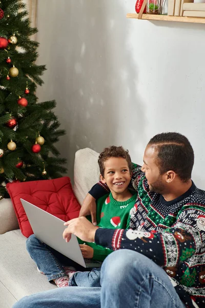 Vertikale Aufnahme eines fröhlichen afrikanisch-amerikanischen Mannes, der seinen Sohn umarmt, während er Weihnachtsfilm auf dem Laptop anschaut — Stockfoto
