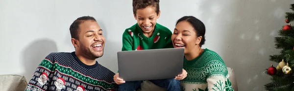 Jolie famille afro-américaine moderne assise sur le canapé regardant un film sur ordinateur portable, Noël, bannière — Photo de stock