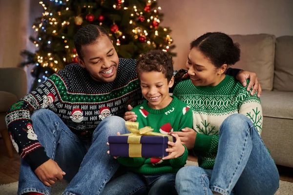 Joyeuse famille afro-américaine en pulls confortables regardant joyeusement son fils tenant présent, Noël — Photo de stock