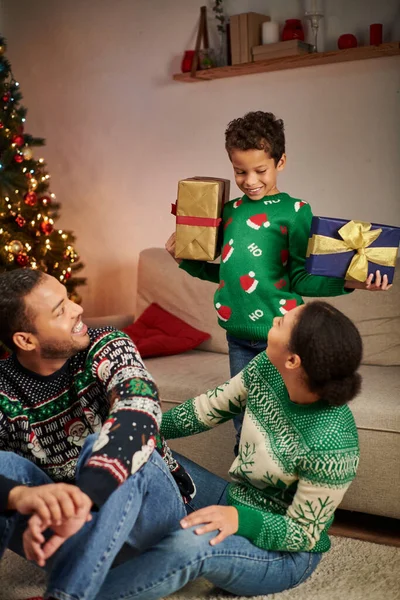 Plan vertical de joyeux garçon afro-américain montrant des cadeaux à ses parents heureux, Noël — Photo de stock