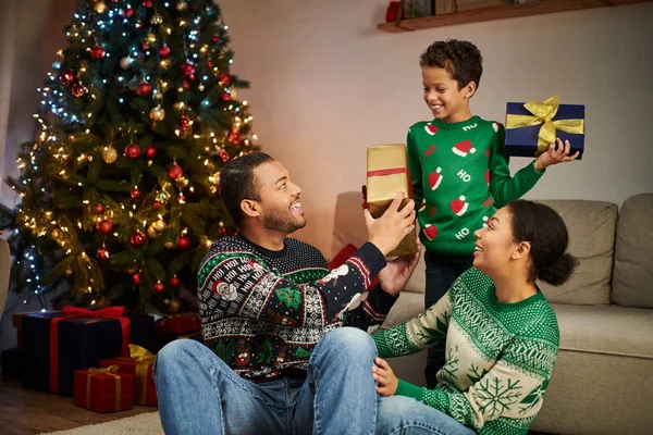 Adorable joyful african american boy showing presents to his parents next to Christmas tree — Stock Photo