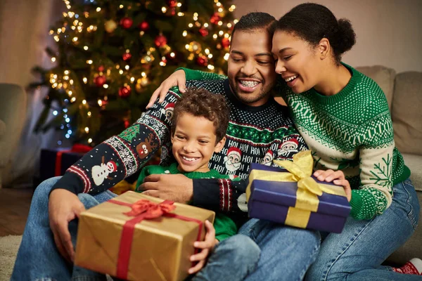 Joyeux parents afro-américains étreignant leur fils alors qu'il tient présent et souriant joyeusement — Photo de stock