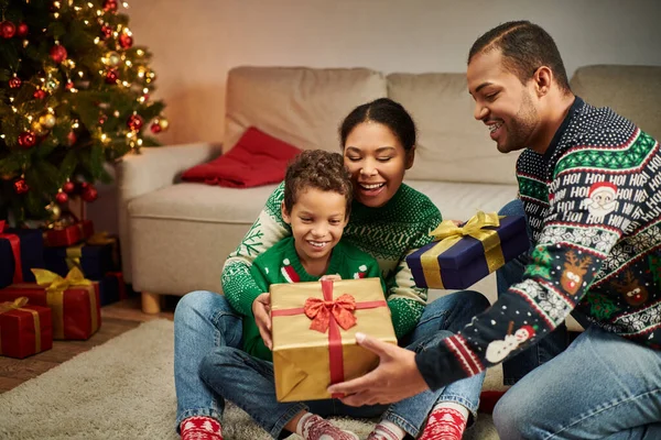 Feliz família afro-americana sentada no chão pela árvore de Natal com guirlandas trocando presentes — Fotografia de Stock