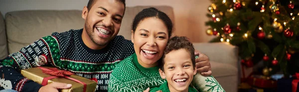 Heureuse famille afro-américaine étreignant chaleureusement et souriant joyeusement à la caméra, Noël, bannière — Photo de stock