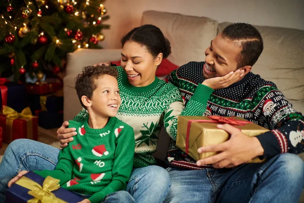 Fröhlicher süßer afrikanisch-amerikanischer Junge mit Geschenk in der Hand und Blick auf seine freudigen Eltern, Weihnachten — Stockfoto