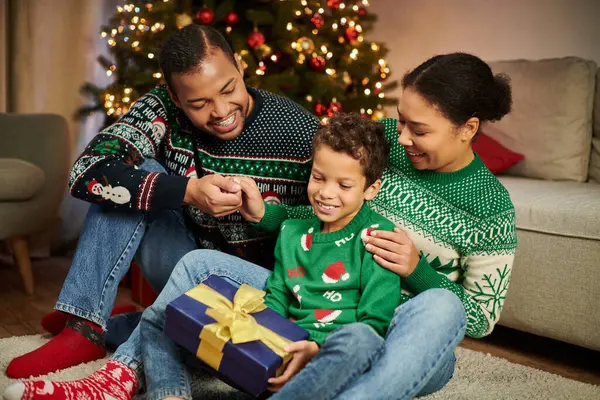 Joyeux parents afro-américains regardant avec amour leur fils alors qu'il tient cadeau de Noël — Photo de stock
