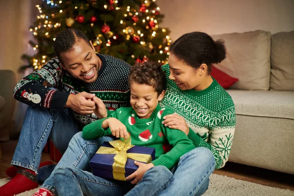 Счастливые африканские американские родители в уютных свитерах смотрят, как их сын распаковывает рождественский подарок — стоковое фото