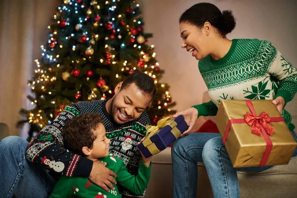 Alegre familia afroamericana en suéteres cálidos sentados junto al árbol de Navidad intercambiando regalos - foto de stock