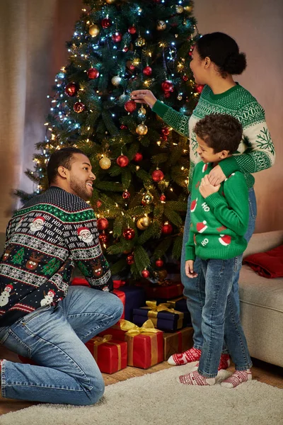 Plano vertical de la familia afroamericana moderna junto al árbol de Navidad sonriéndose entre sí - foto de stock