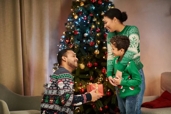 Bom olhar afro-americano alegre homem segurando presente e olhando para sua esposa e filho, Natal — Fotografia de Stock