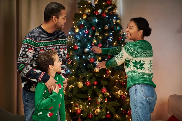 Mujer afroamericana feliz decorando el árbol de Navidad mientras su hijo y su marido le sonríen - foto de stock