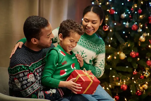 Alegre lindo africano americano chico sosteniendo presente mientras sus padres sonriendo a él, Navidad - foto de stock