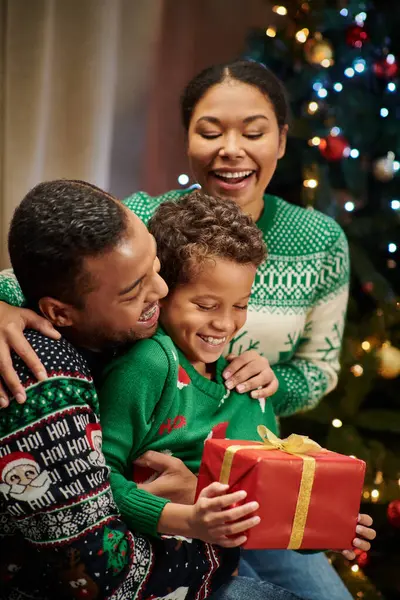 Vertical shot of adorable african american boy holding present surrounded by his jolly parents — Stock Photo